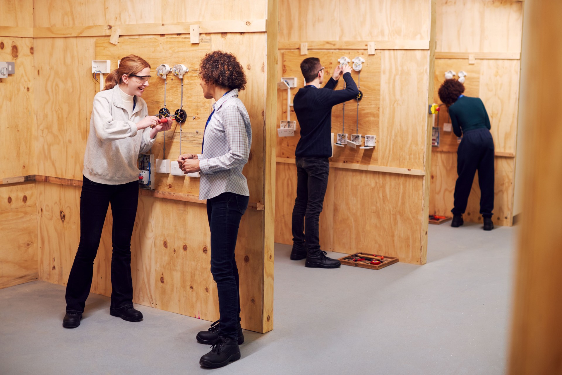 Female Tutor With Trainee Electricians In Workshop Studying For Apprenticeship At College